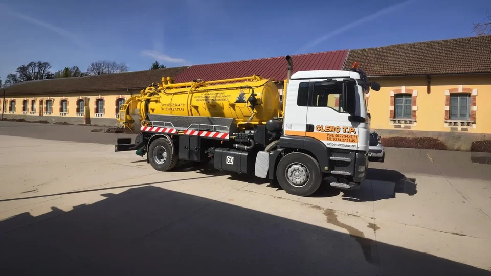 Camion d'assainissement jaune stationné devant un bâtiment historique en pierre, tuyaux verts déployés vers une bouche d'égout pour vidange ou curage